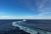 A naval ship sails to the right in the ocean during a clear day with blue sky, leaving ripples of white in its wake.