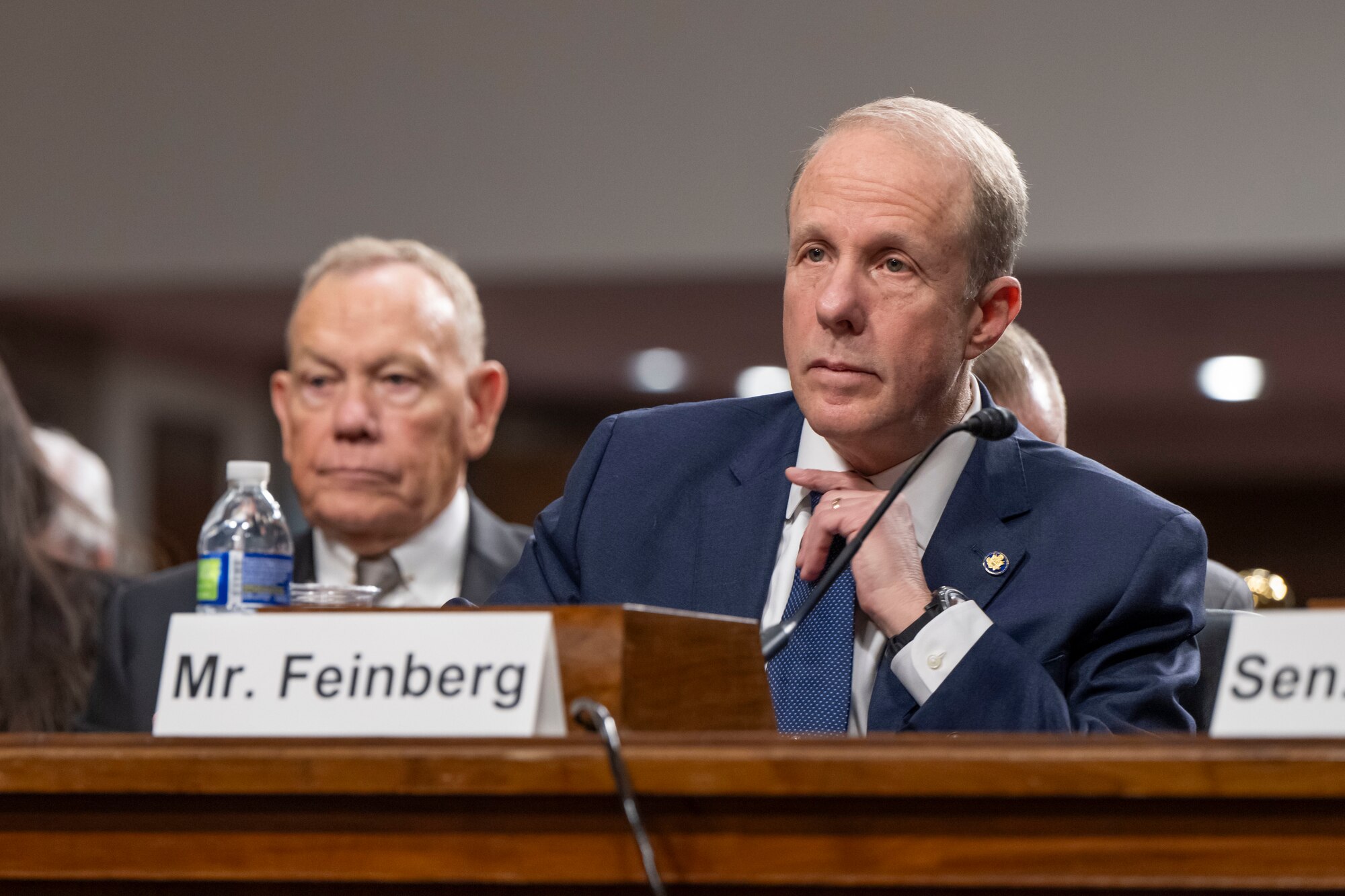 A civilian sits by a microphone at a table, with another person seated in the background.