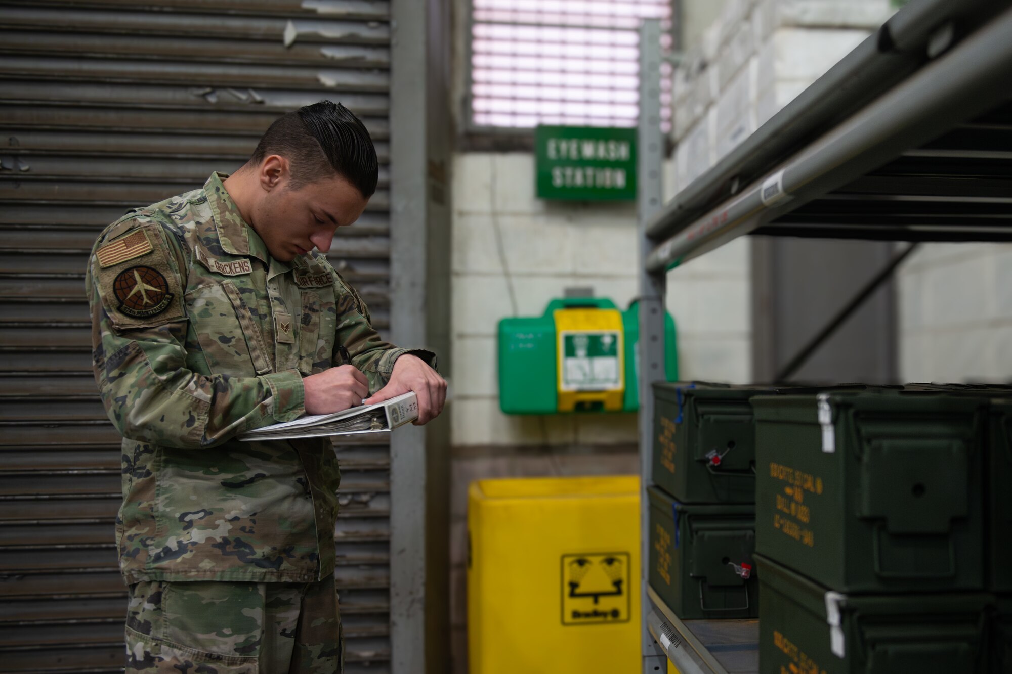 An Airman stands with a clipboard and pen in hand checking green ammo boxes on shelves.