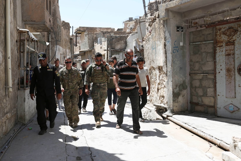 Men in uniforms walk down a narrow street with boys from the neighborhood.