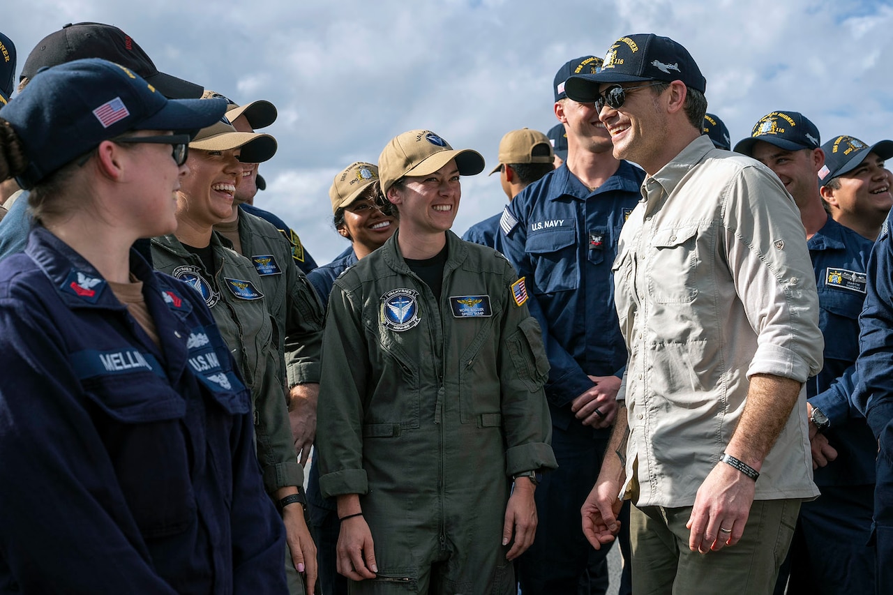 Defense Secretary Pete Hegseth smiles while talking with smiling sailors standing outdoors.
