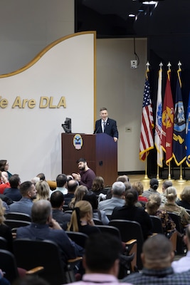A man in a suit stands at a podium and speaks to a large audience in an auditorium.