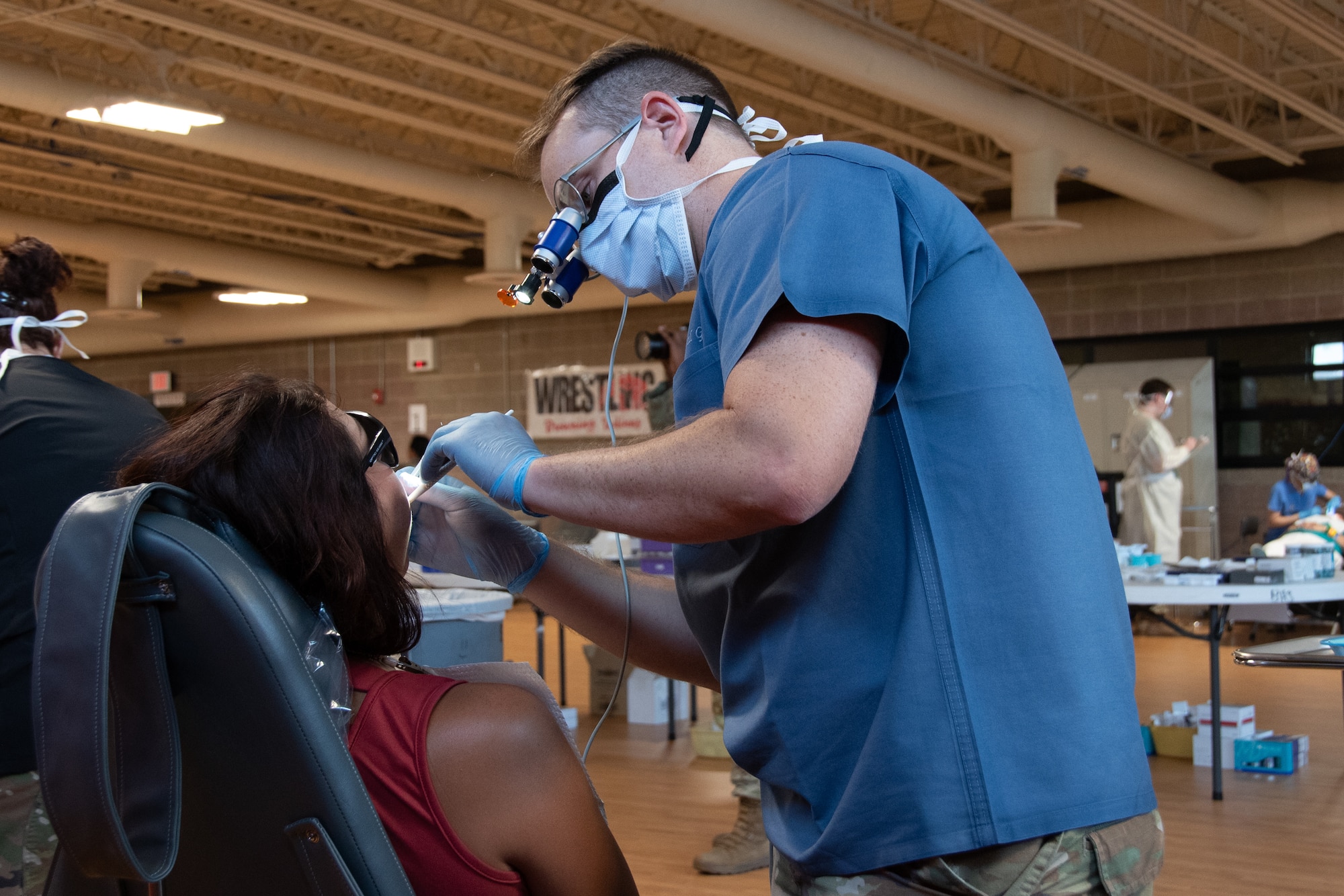 An Air Force dentist in blue scrubs examines a seated civilian's teeth while wearing a medical mask, plastic gloves, and a dentistry loupes.