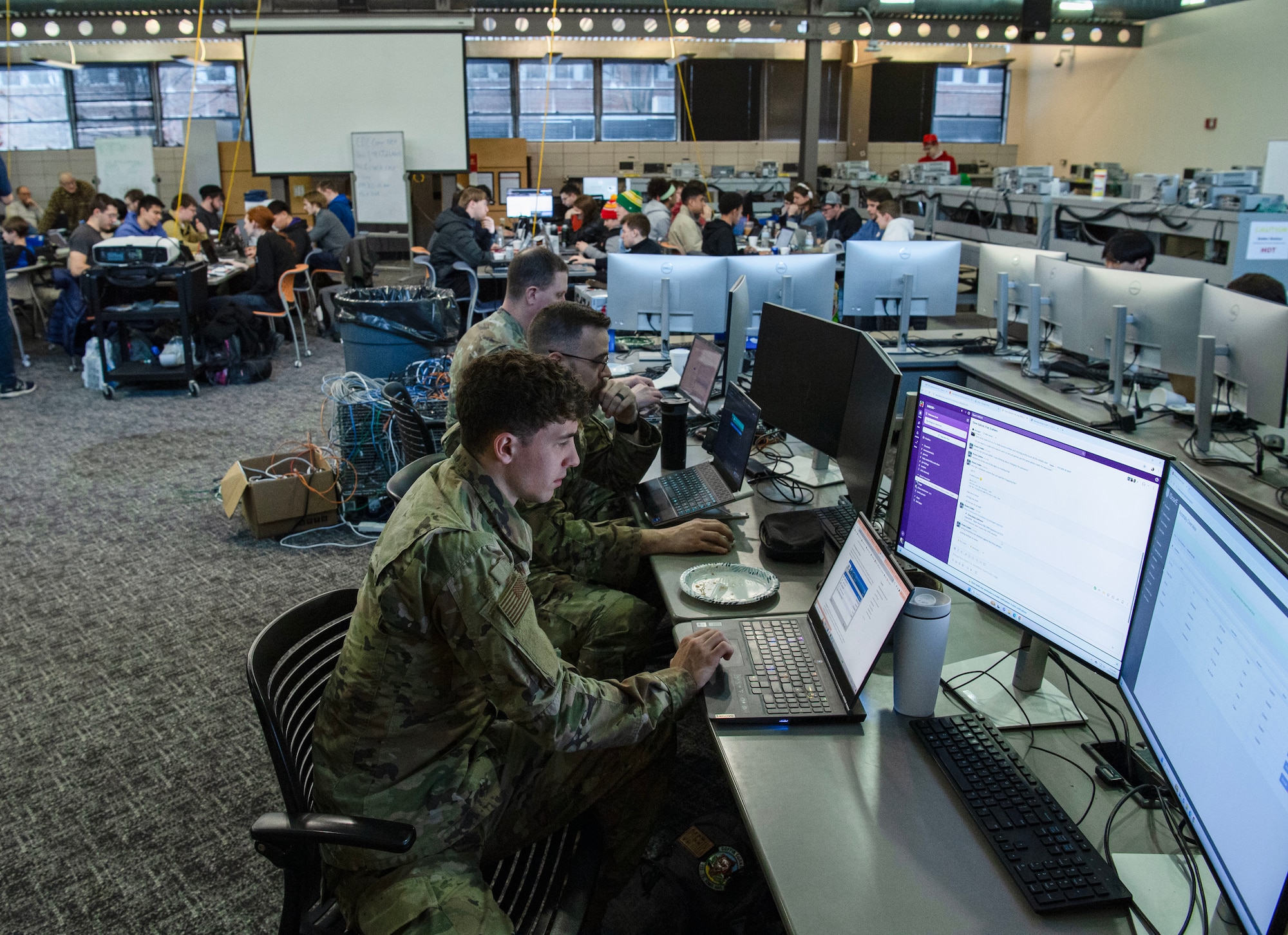 Airmen in uniform type on computers in a large classroom with dozens of civilian students