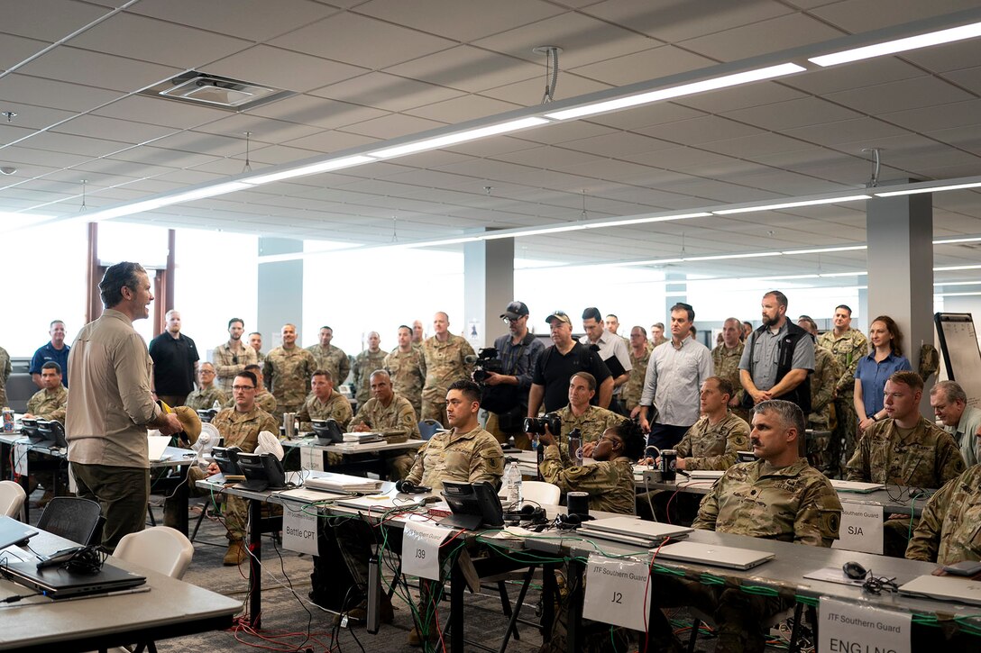 A government official speaks in front of troops seated at tables in a classroom.