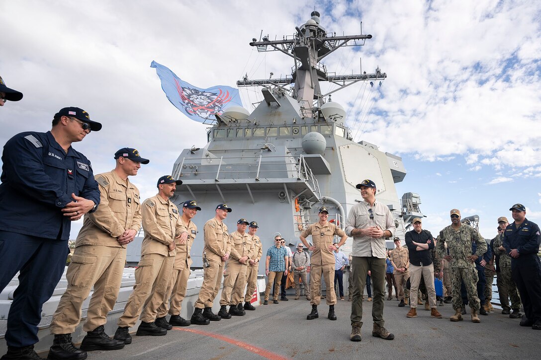 A civilian stands in front of a line of sailors on the flight line of a military naval ship.