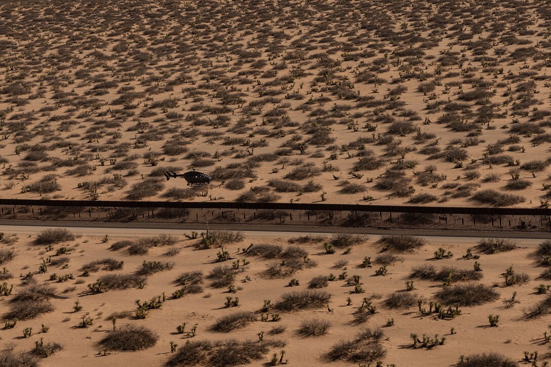 A helicopter flies low close to a wall cutting through a desert.