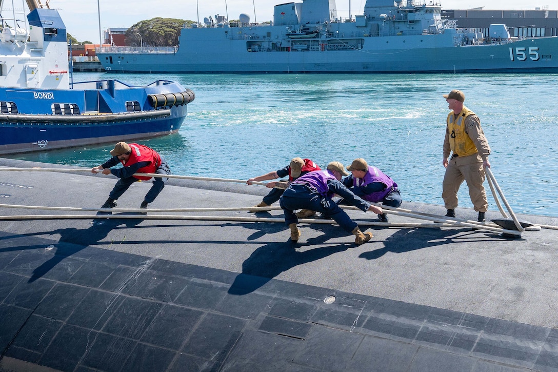 A sailor shouts as fellow sailors in colorful vests pull lines on top of a submarine on a sunny day with ships in the background.