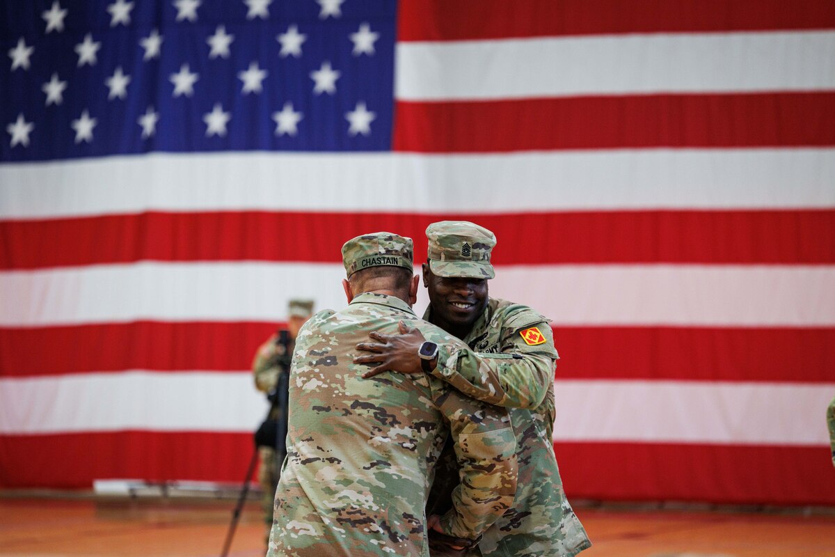 Two soldiers hug in front of a large American flag while another stands in the background.
