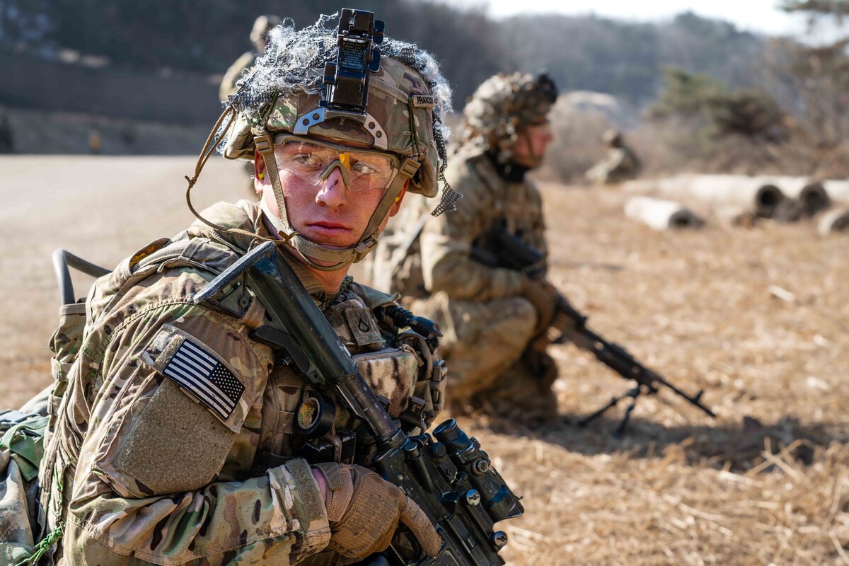Two soldiers with weapons and gear kneel on a field as mountains loom in the background.