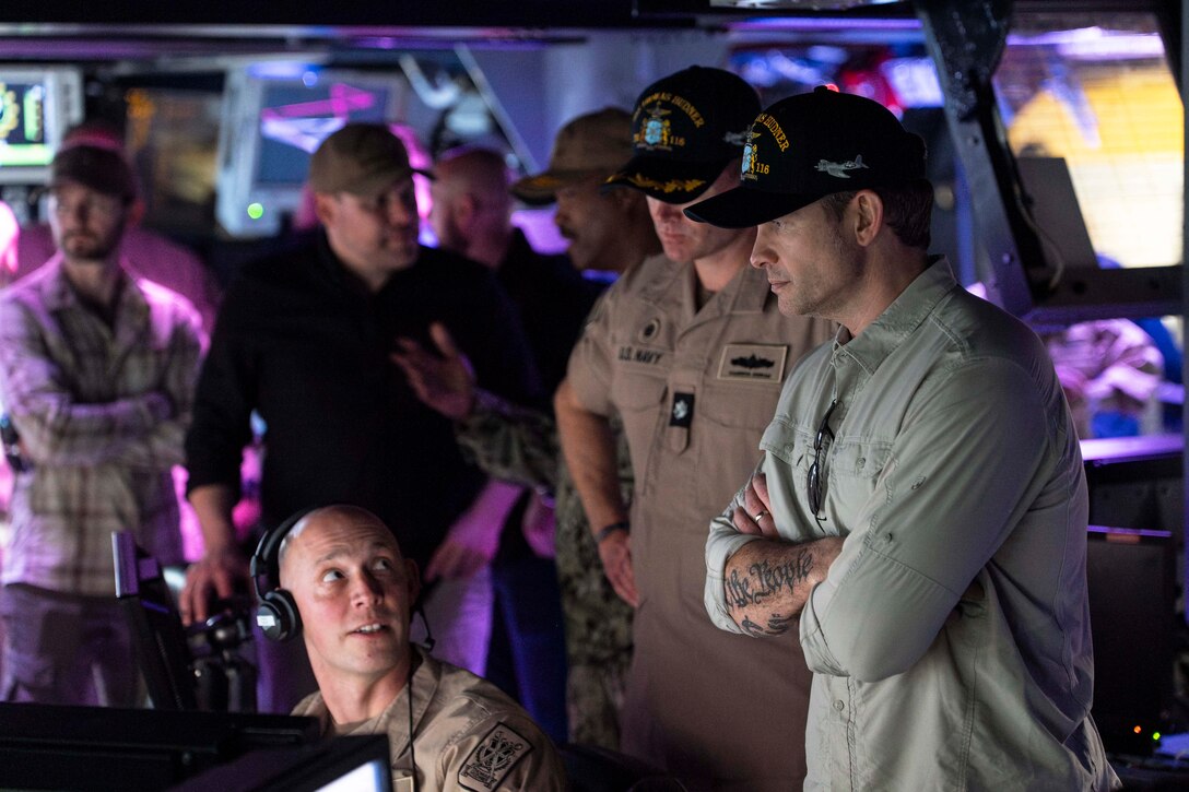 The defense secretary stands with sailors while looking at a monitor in a dimly lit control room aboard a ship illuminated by purple light.
