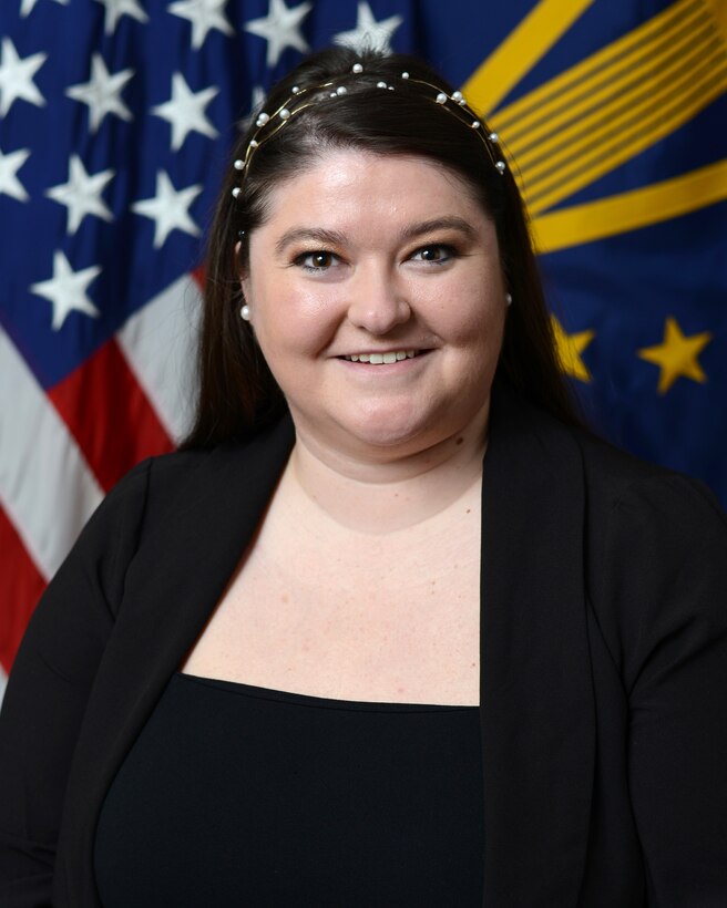 A woman in business attire sits in front of two flags.