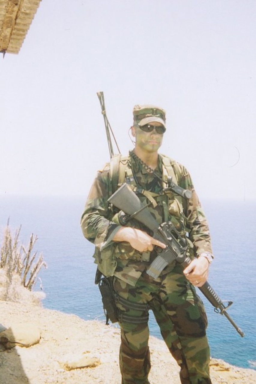 A man in a camouflage uniform with sunglasses and camouflage face paint is holding a rifle while posing for the camera.