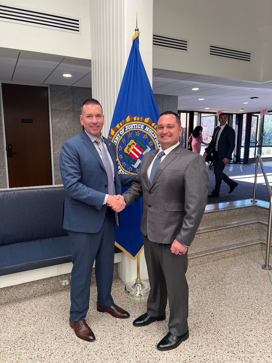 Two men shake hands in front of a flag