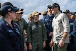 Defense Secretary Pete Hegseth smiles while talking with smiling sailors standing outdoors.