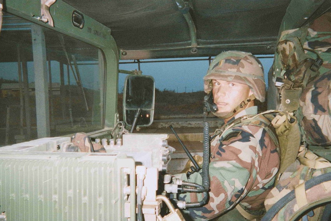 A man in a camouflage uniform is looking at the camera while wearing a helmet and talking on a phone inside a military vehicle.