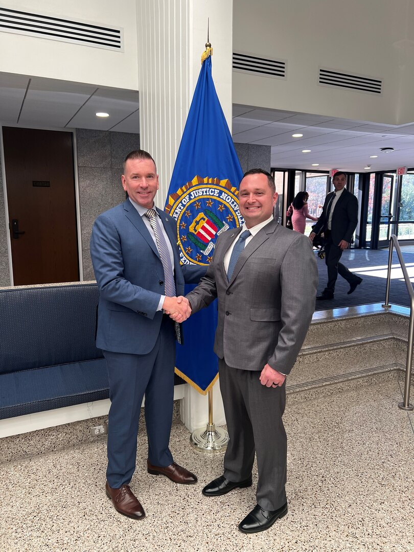Two men shake hands in front of a flag