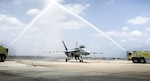 Photo of F/A-18 Super Hornet on a flight line with two fire trucks on either side spraying water over it in a water salute.