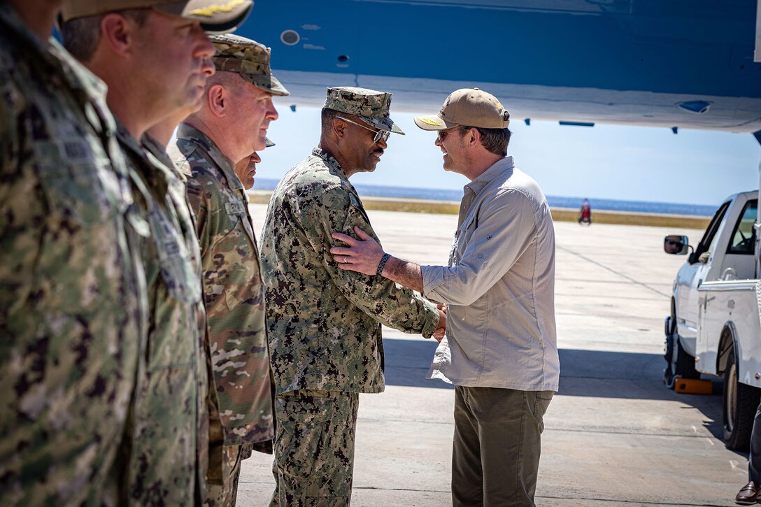 A man in civilian attire greets a sailor among a line of sailors on the tarmac of an air field.