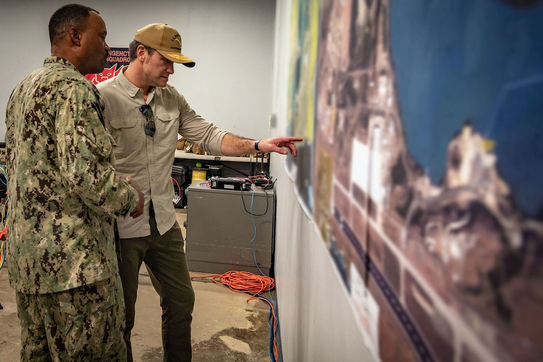 A government official in civilian attire points to a blurred out poster on a wall while a sailor looks on.