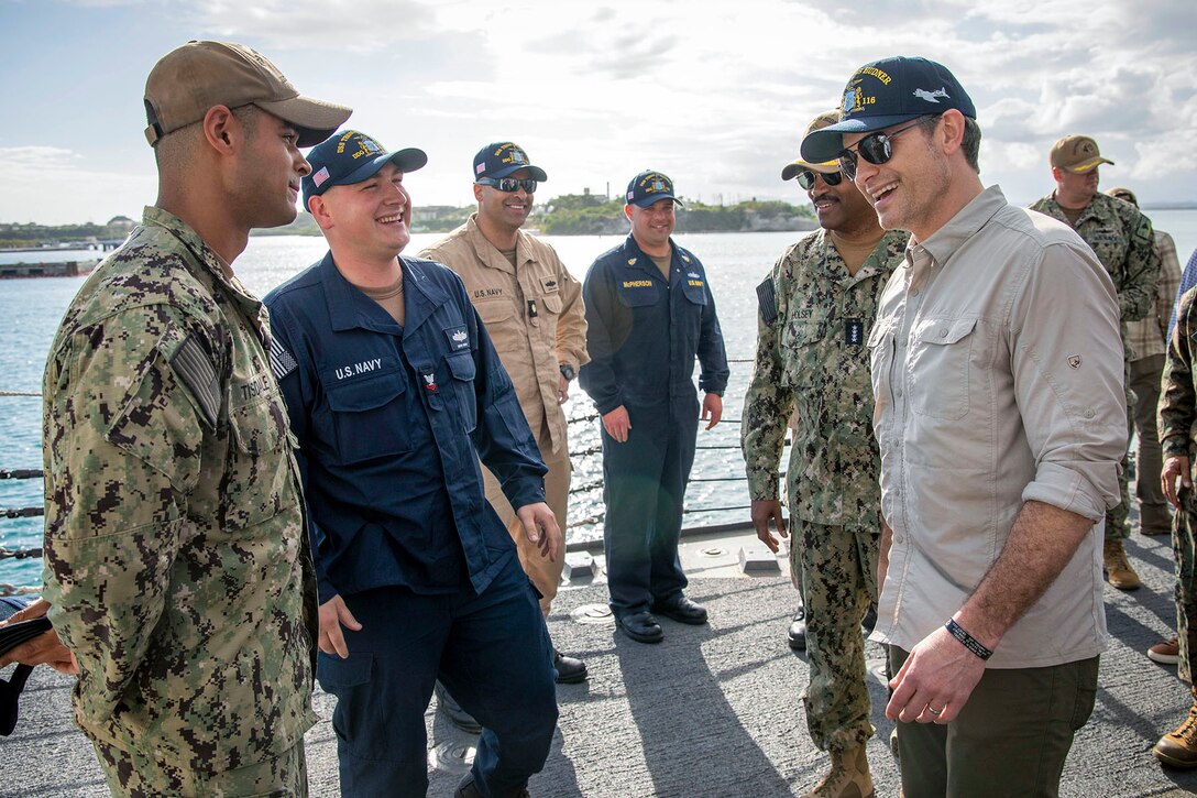 A government official in civilian attire greets sailors on the deck of a naval military ship during the day.