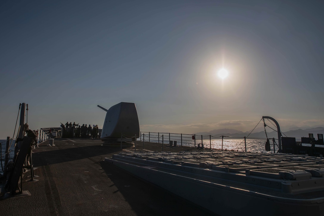 Sailors gather in the front of a ship at sea near a weapon as the sun beams down with mountain silhouettes in the distance.