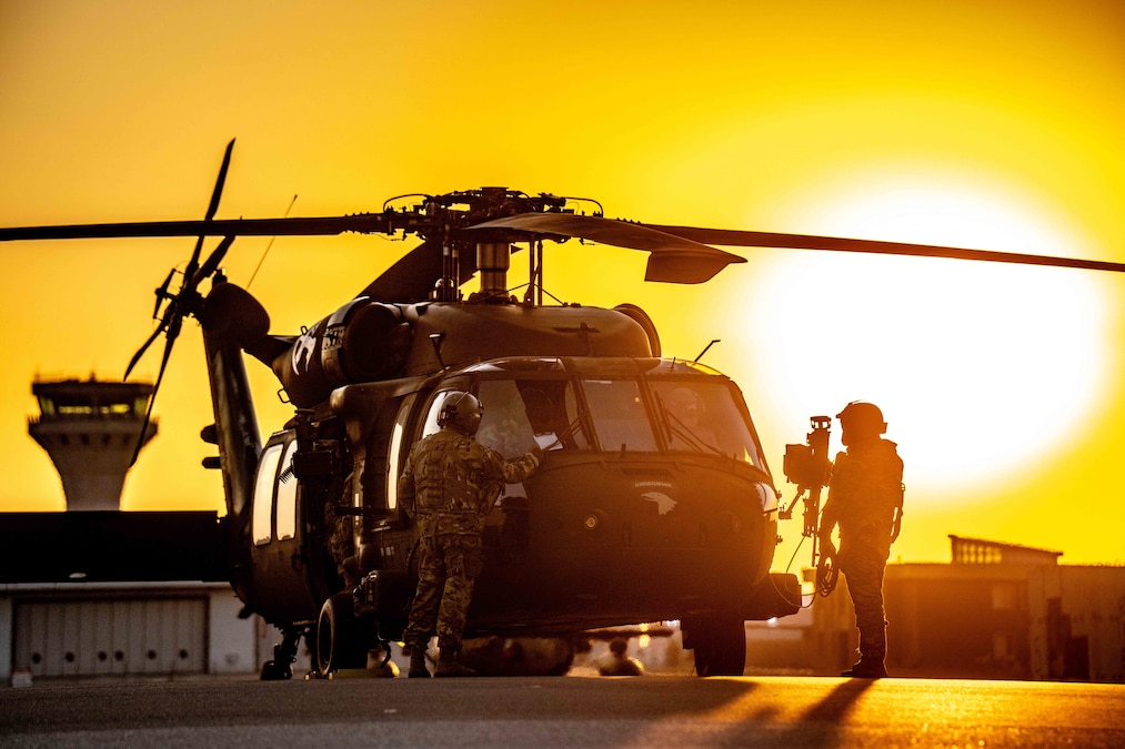 Two soldiers stand by a helicopter parked at an air base with the sun in the background emitting an orange hue.