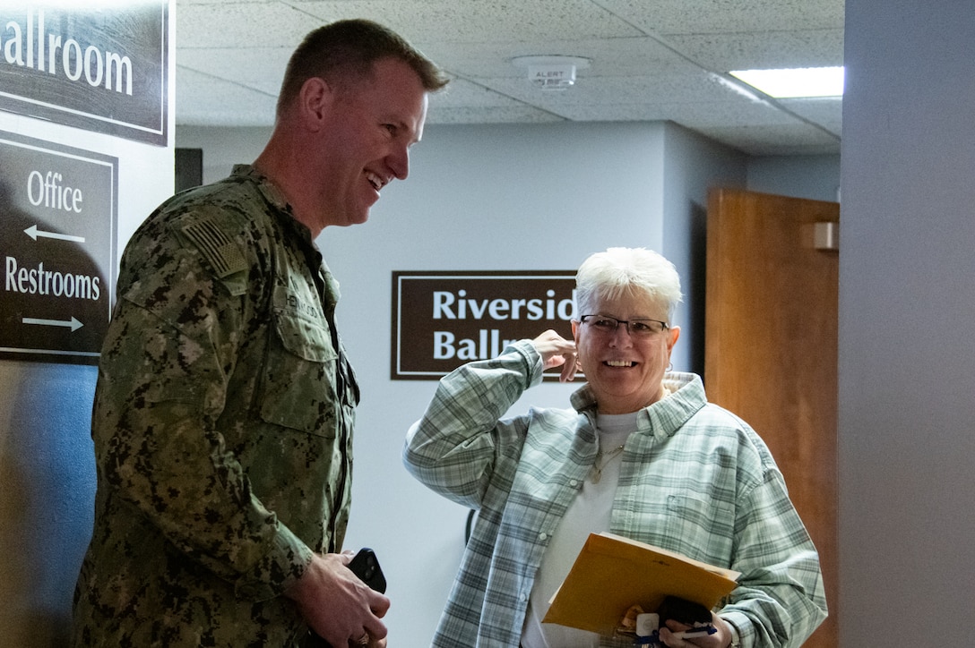 A man in a camouflage military uniform smiles at a woman in a hallway.