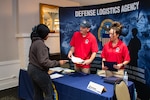 A person talks to two other people standing behind a table at an information booth.