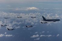One refueler flies in front of 7 fighter jets over the clouds. There is a mountain in the background.
