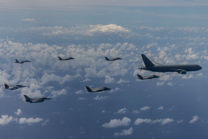 One refueler flies in front of 7 fighter jets over the clouds. There is a mountain in the background.