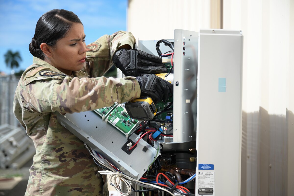 An airman wearing protective gloves uses a drill while working outdoors on a circuit board on a sunny day.