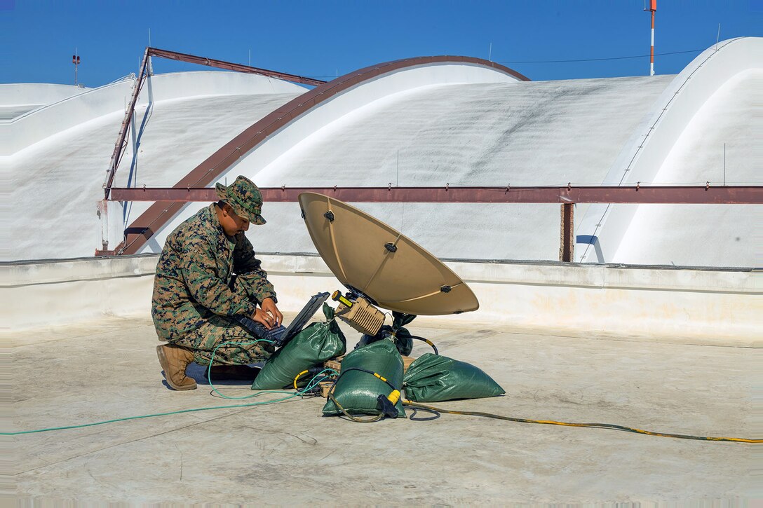 A Marine kneels to check satellite connections on top of a building.