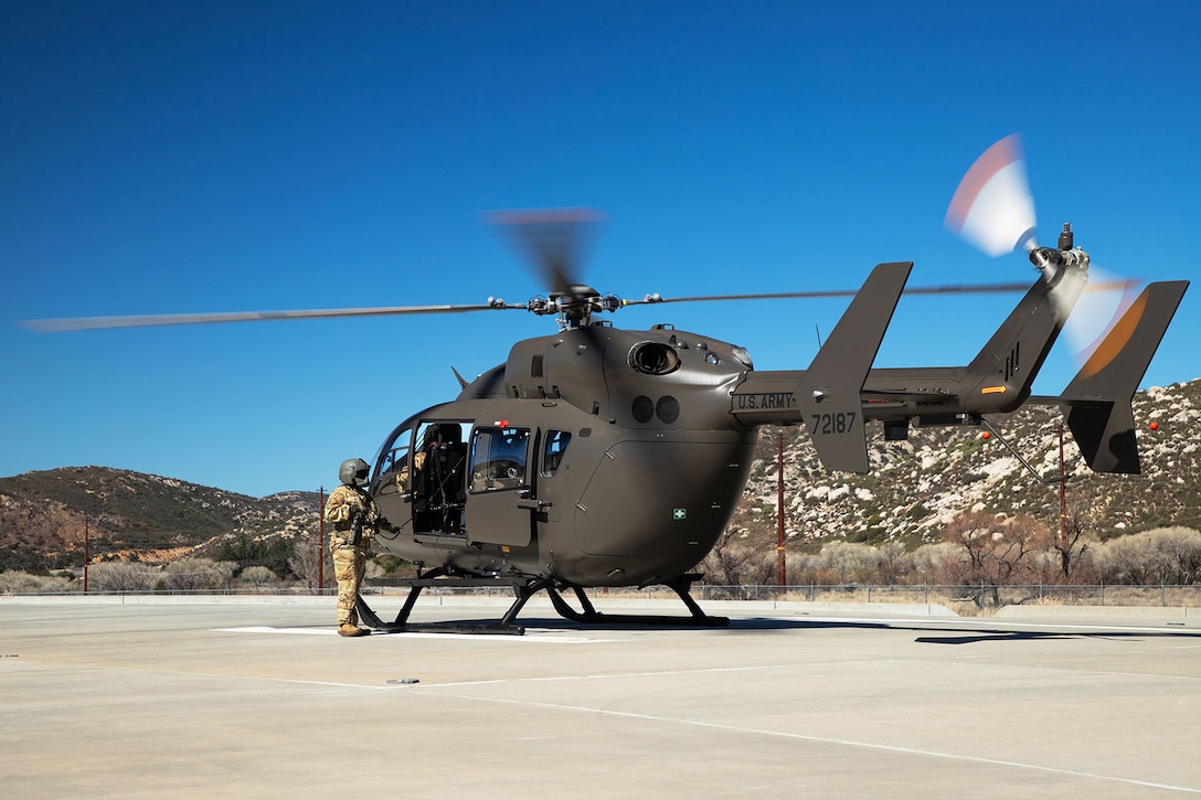 Soldiers work on a helicopter on a tarmac with mountains in the background under a blue sky.