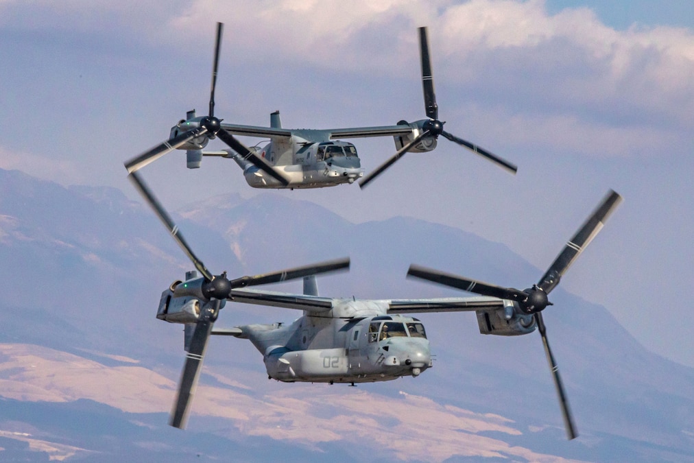 Two military aircrafts fly closely together in a cloudy sky above a sloped, mountainous terrain.