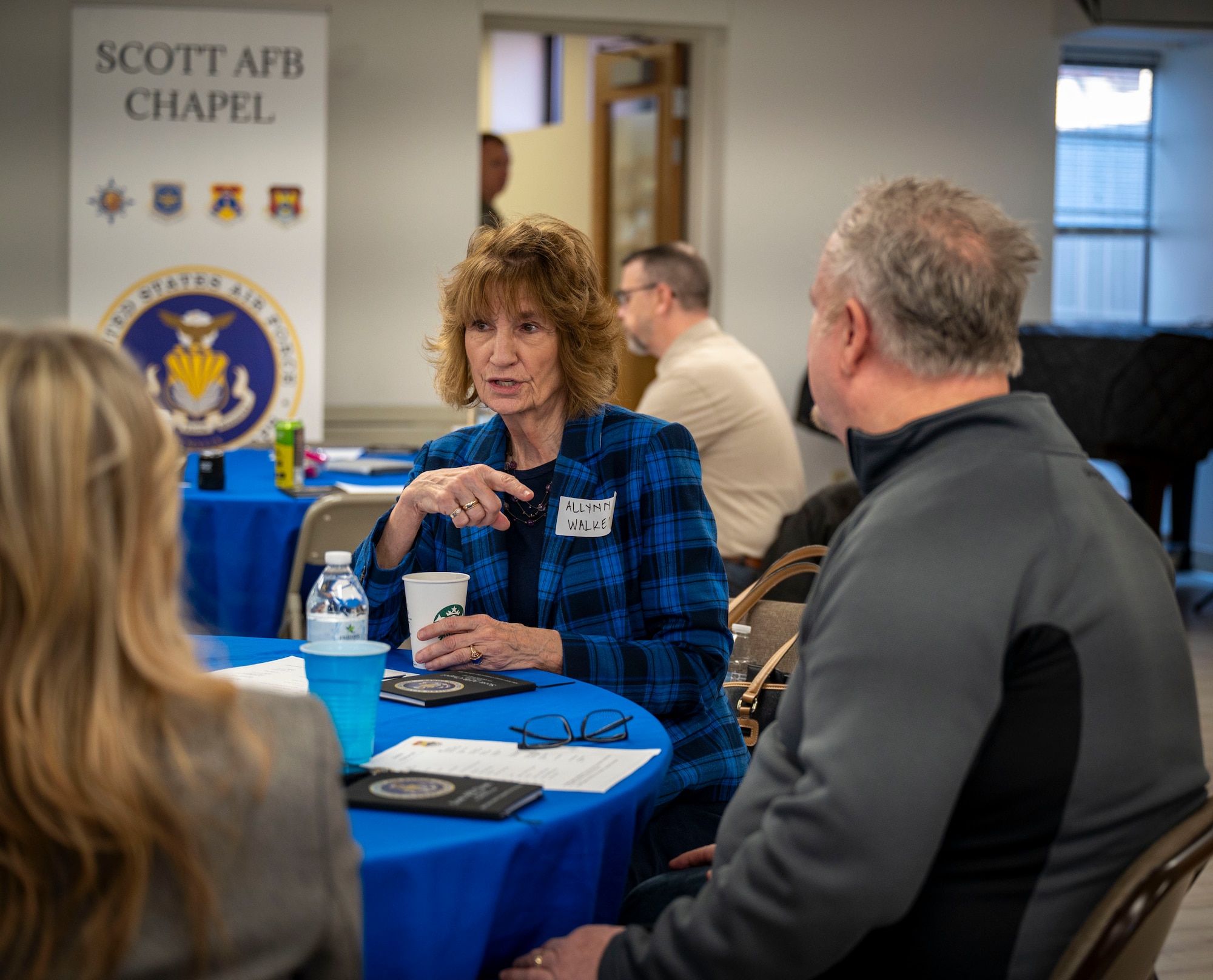 Rev. Allynn Walker, wearing a blue and black checkered blazer converses with others seated at her table.