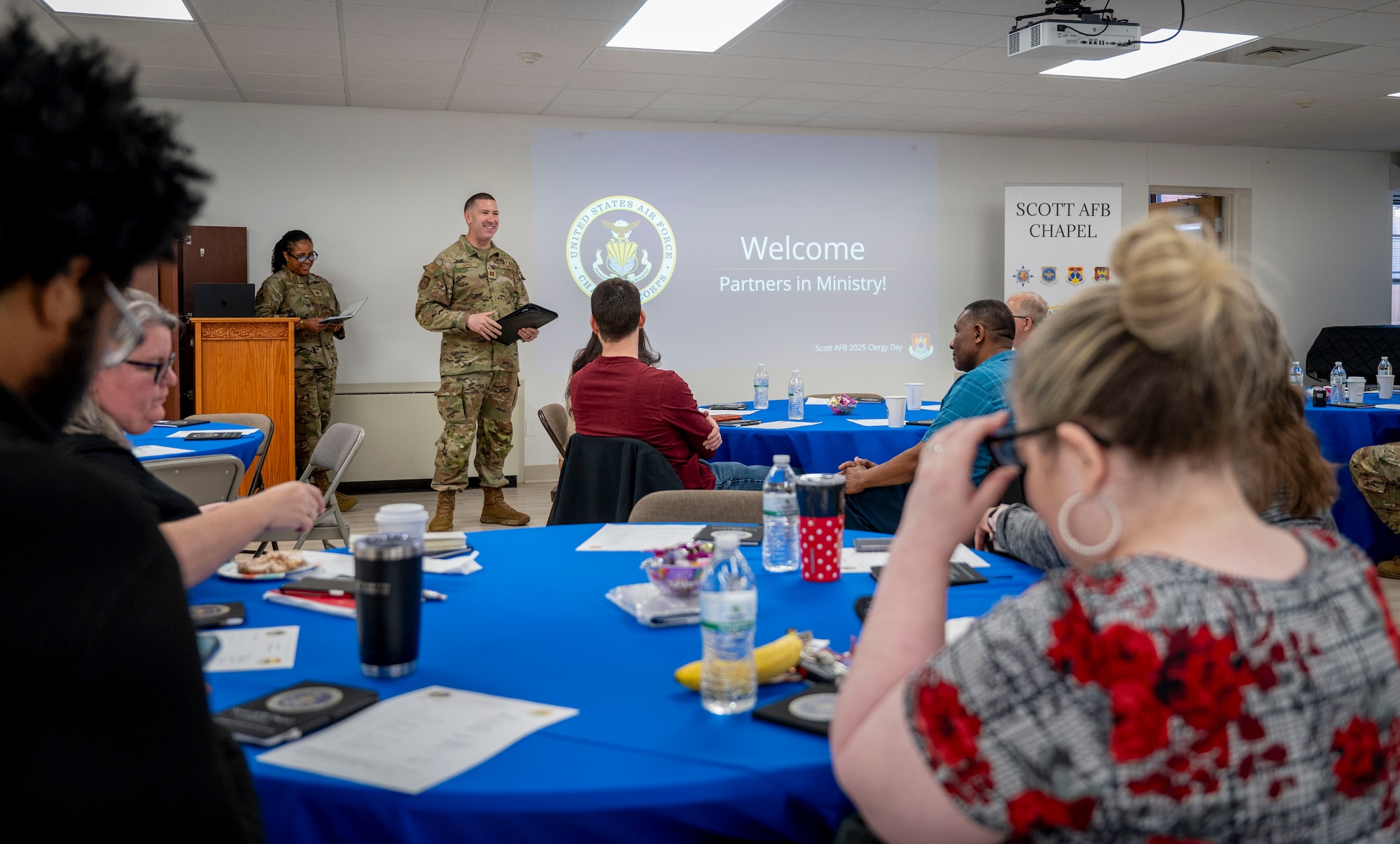 Two chaplains stand at the front of a room welcoming attendees to the annual clergy day event.