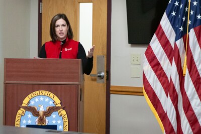 A woman in a red vest speaks behind a lectern with the Defense Logistics Agency logo on it.
