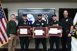 A group of five people pose for a group photo. The three in the center of the group are wearing police uniforms and holding framed award certificates.