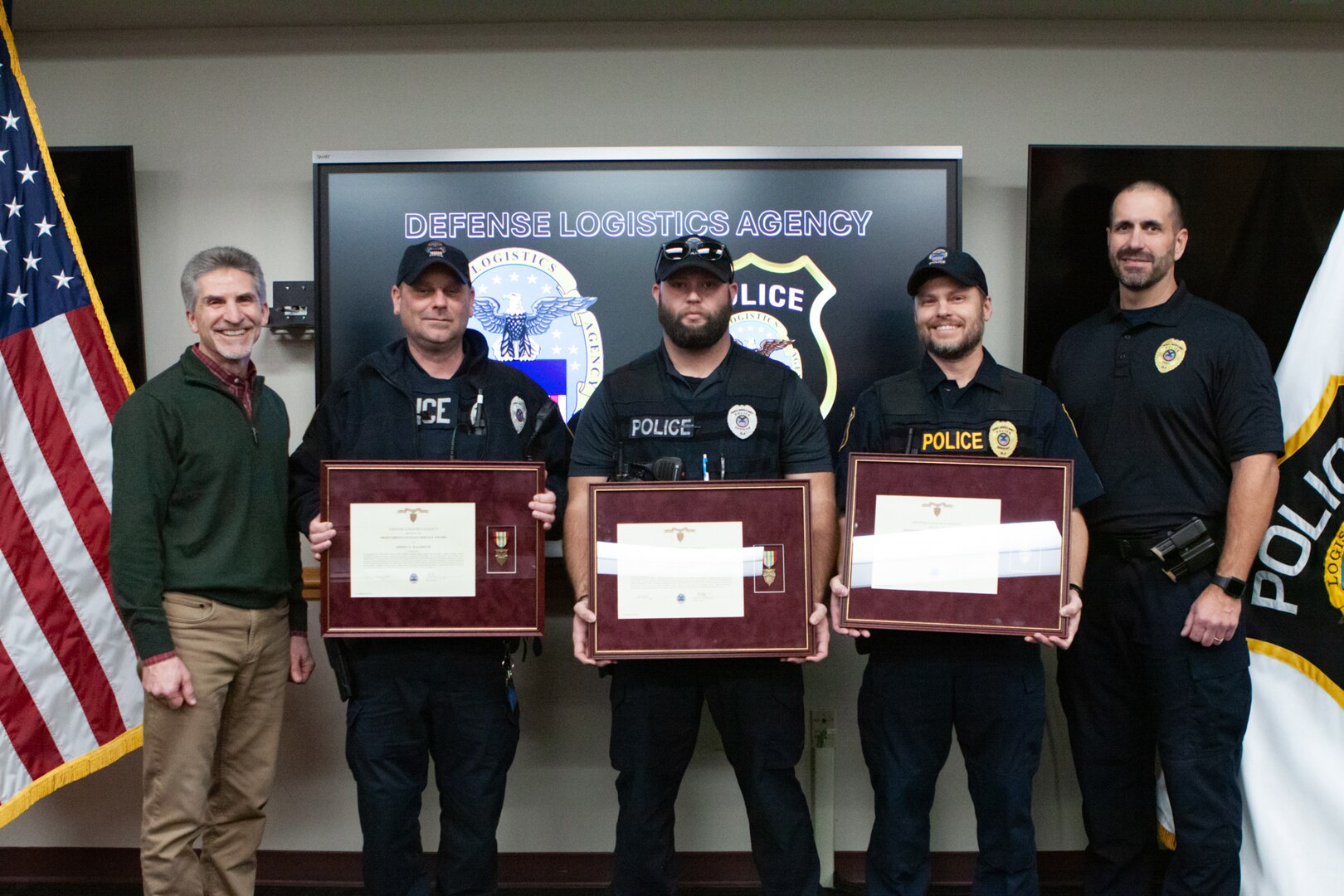 A group of five people pose for a group photo. The three in the center of the group are wearing police uniforms and holding framed award certificates.