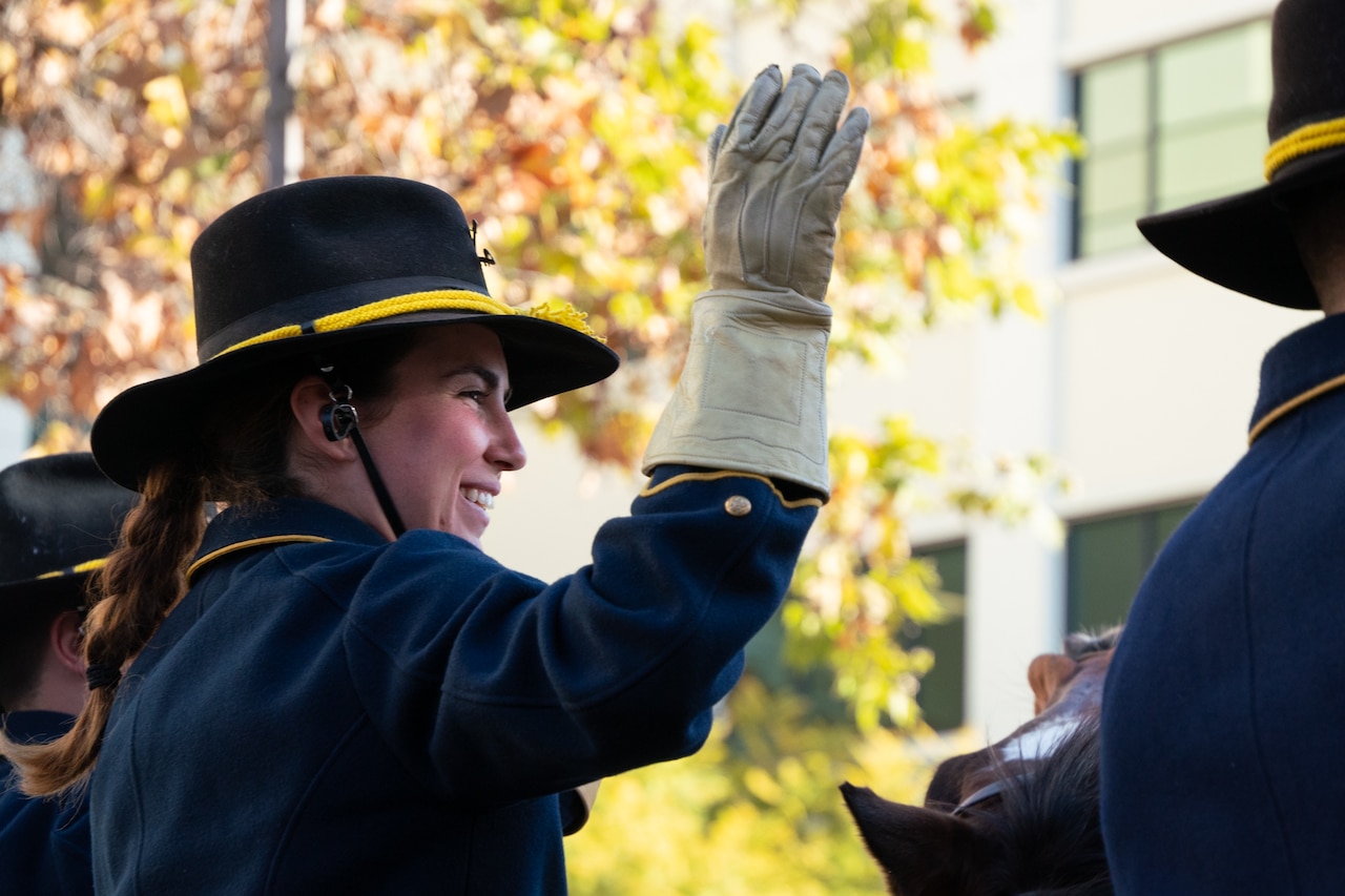 A soldier wearing a glove and hat waves and smiles.