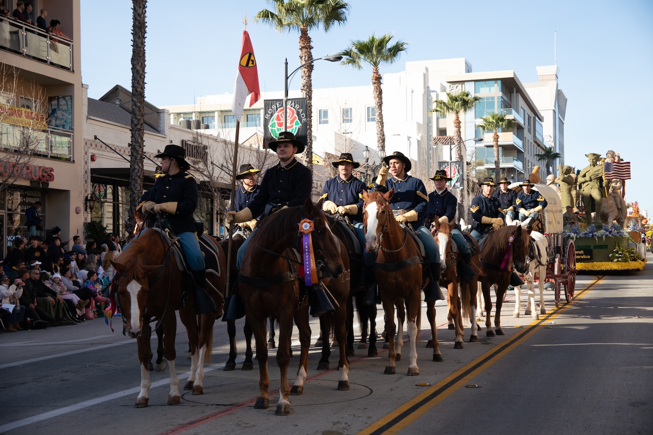 A group of mounted soldiers move down a parade street lined with buildings and palm trees.