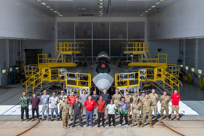 A large group of airmen and civilians stand in front of yellow stands in a hangar.