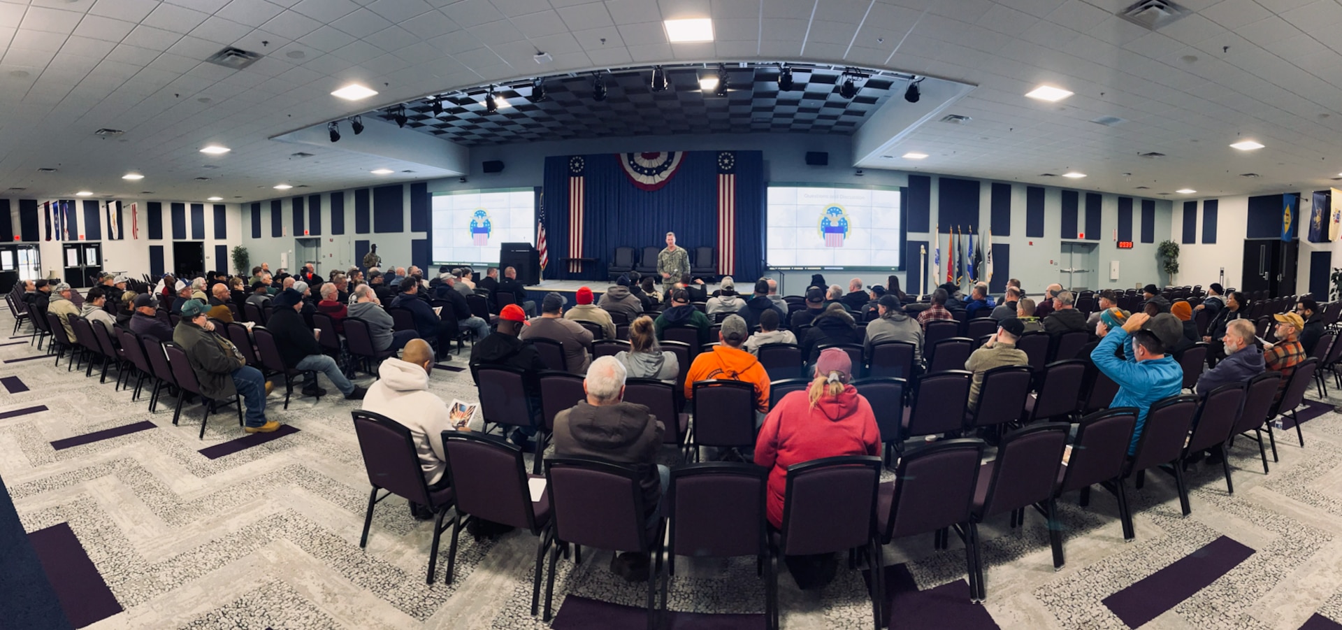 A large group of people seated in a large auditorium look toward a man in a camouflage military uniform speaking at the front of the room with two digital screens behind him.