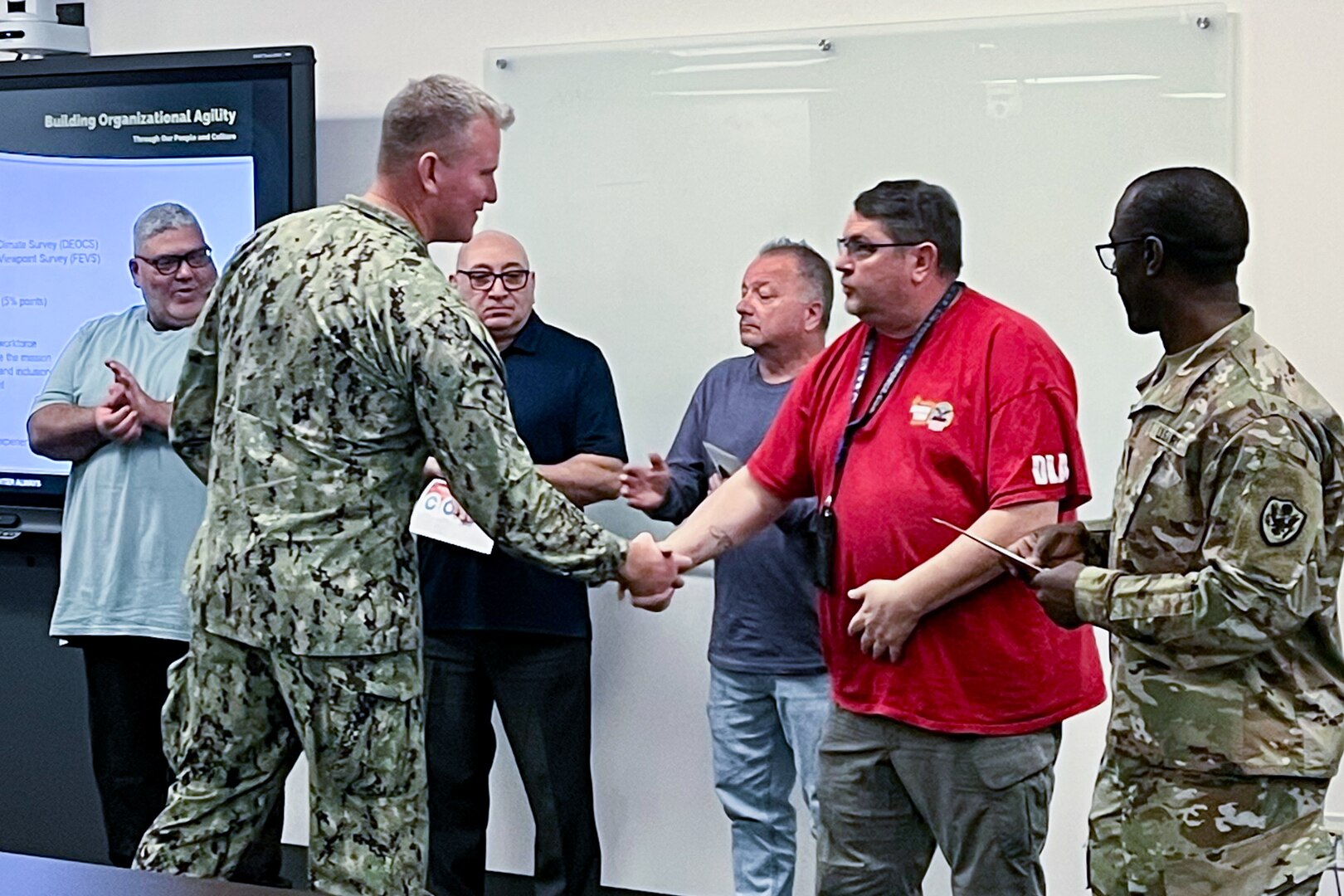 A man in a camouflage military uniform shakes another man's hand with people clapping in the background.