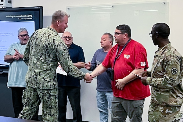 A man in a camouflage military uniform shakes another man's hand with people clapping in the background.