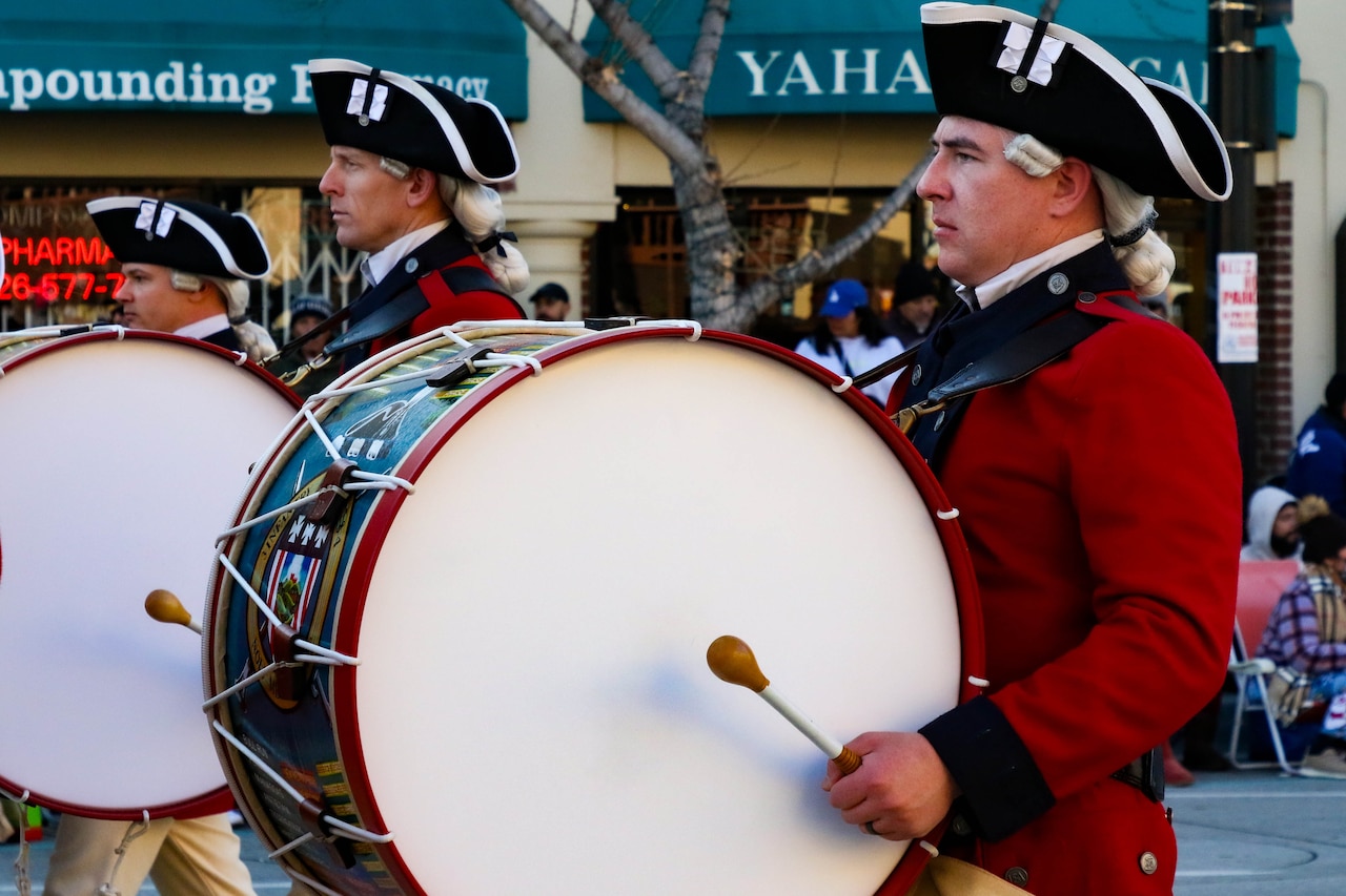 A line of drummers in Revolutionary era military attire march down a road.