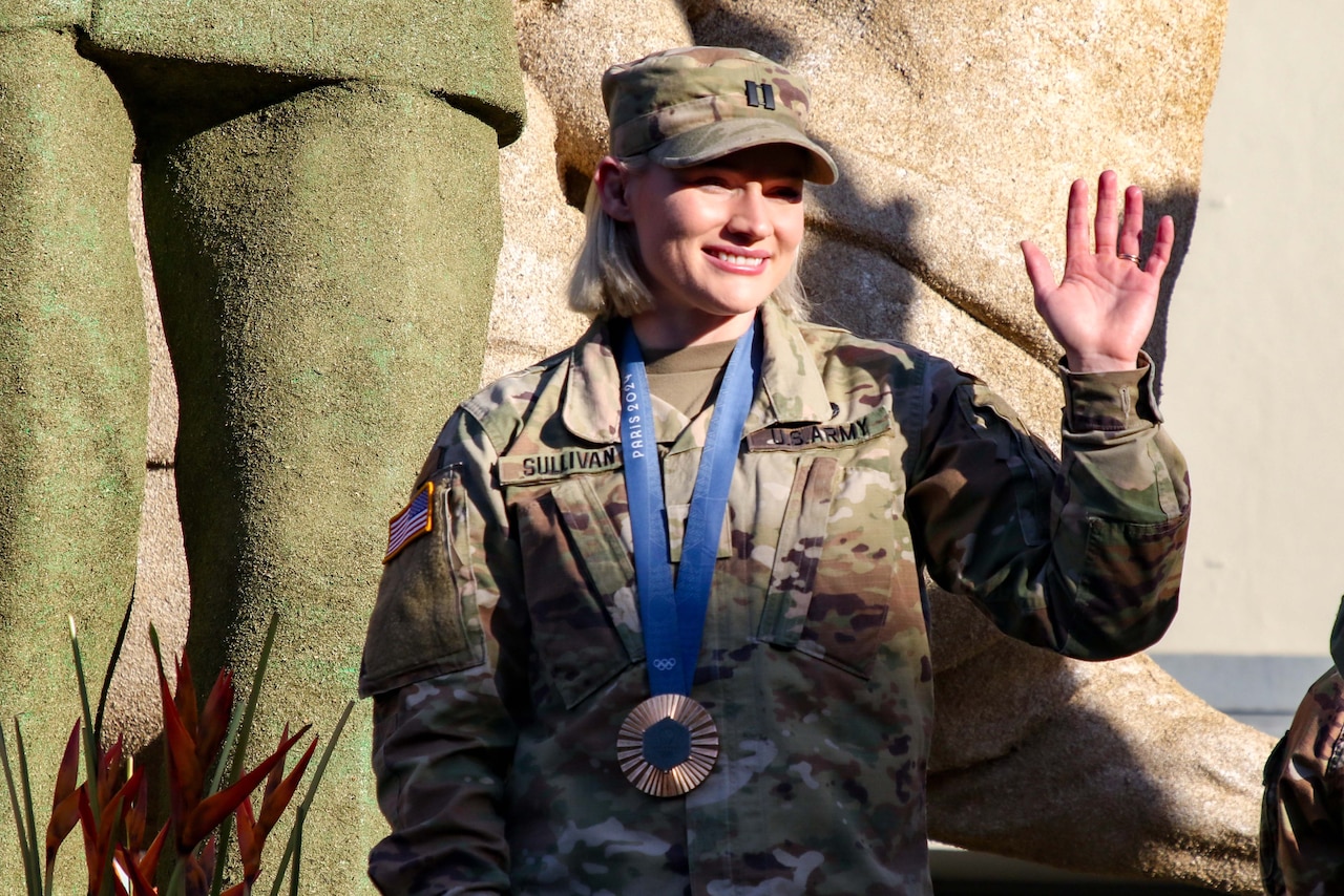 A soldier with an Olympic bronze medal waves in front of statues.