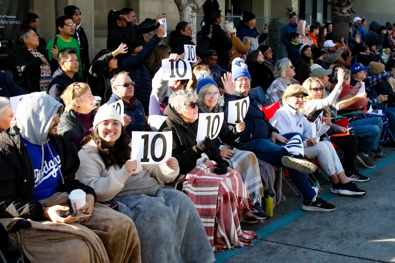 Spectators of the 2025 Army Rose Parade float hold up score cards and sit by a road.