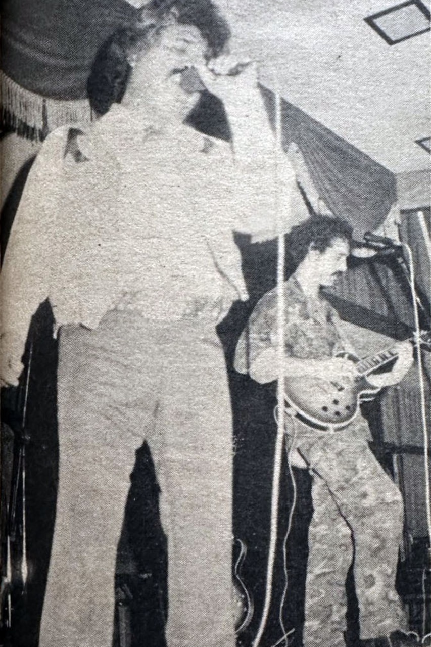 A man sings into a microphone and another strums the guitar on stage.